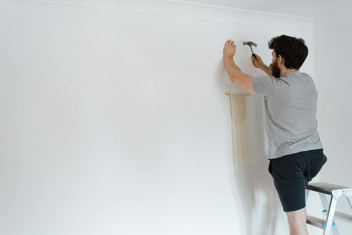 Back view of unrecognizable unshaven male in casual wear using hammer while putting decoration on wall at home