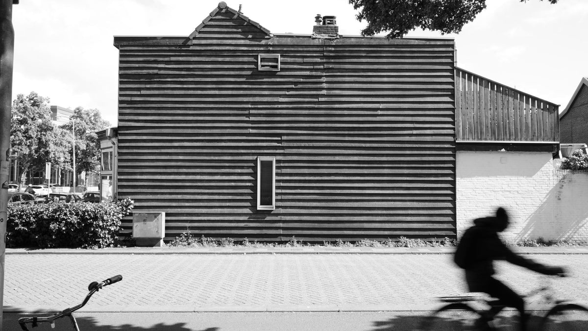 Black and white image of a cyclist's silhouette against a wooden building, capturing motion.