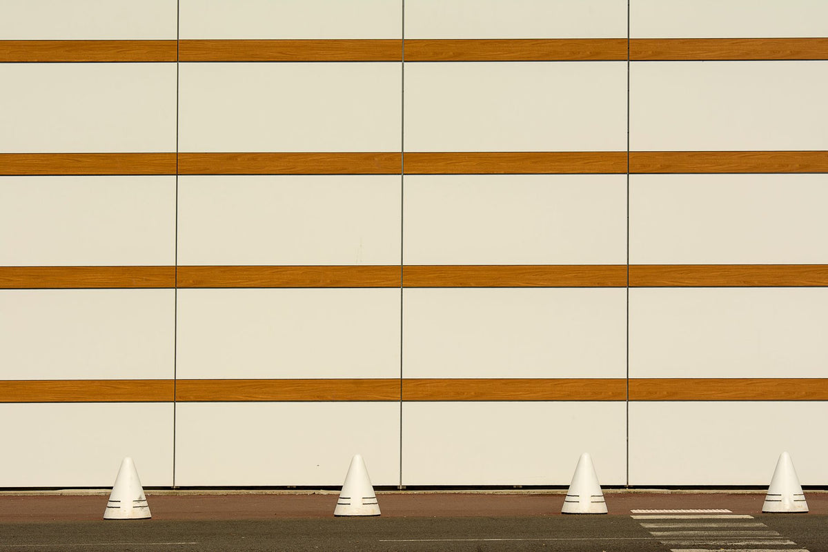 A minimalist architectural wall with horizontal stripes and cones on a sidewalk.