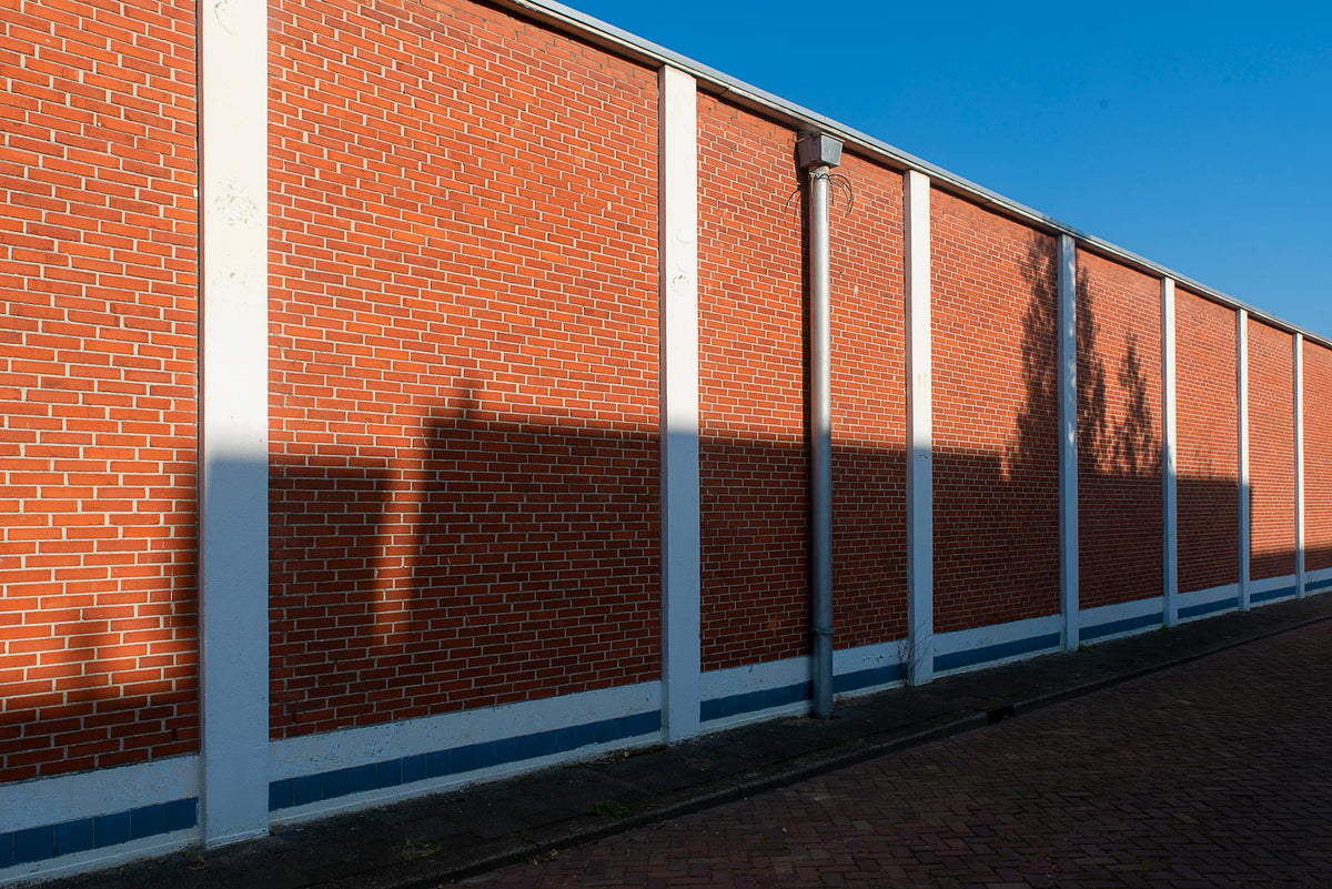 A long red brick wall with geometric shadows cast under clear blue sky.